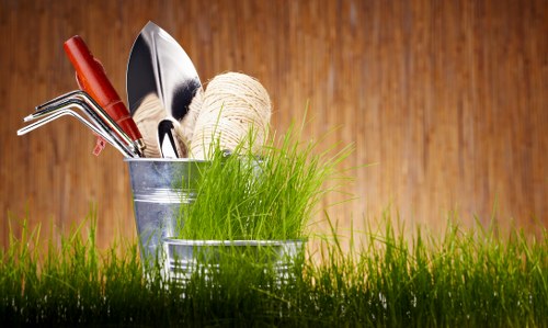 The image features a close-up view of gardening and landscaping tools placed on a vibrant, well-maintained lawn in front of a wooden background. In the foreground, short, lush green grass covers the ground, suggesting a healthy and neatly kept outdoor space typical of residential gardens in Chelsea. Three small metal containers hold various gardening tools, including a trowel, a garden fork, and a pair of hedge shears, along with a ball of twine and a bunch of fresh, upright green grass or plants. The background consists of a warm-toned wooden surface, evoking a natural and inviting garden environment, suitable for outdoor maintenance and lawn care services as offered by Lawn Mowing Chelsea. The overall scene implies readiness for garden upkeep, planting, or general landscaping activities in a UK garden setting, with natural lighting that highlights the textures of the tools, grass, and wood, emphasizing a professional and tidy outdoor environment.